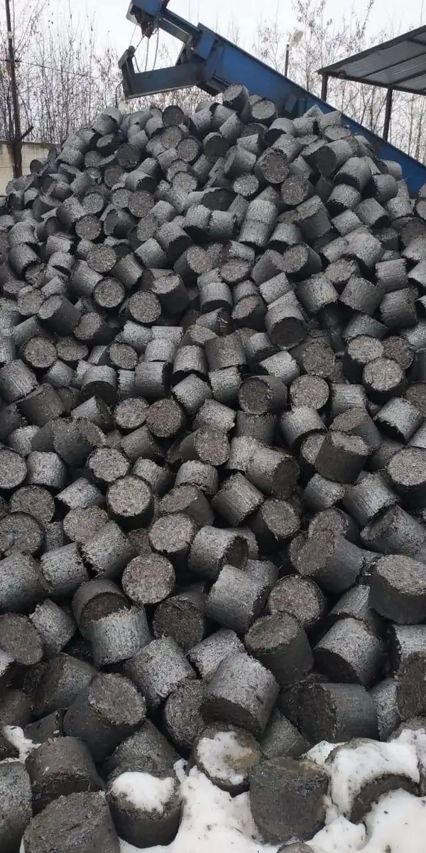 Outdoor pile of cylindrical metal briquettes beneath a blue discharge conveyor.