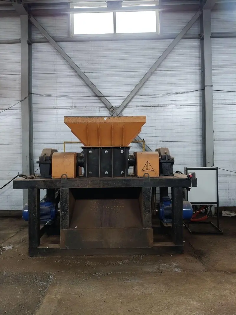 Double-shaft metal shredder with an orange feed hopper and two drive motors, standing on a steel frame inside the plant.