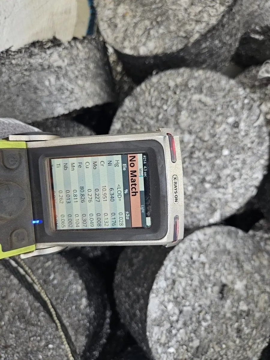 Handheld XRF analyser resting on a stack of metal briquettes, its screen showing an elemental readout and a No Match result.